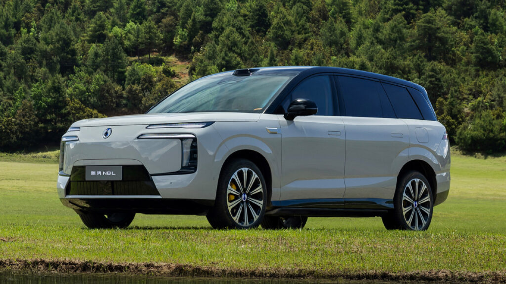 A silver Denza N8L flash charging PHEV parked on grass, shown from the front three-quarter angle against a wooded backdrop.