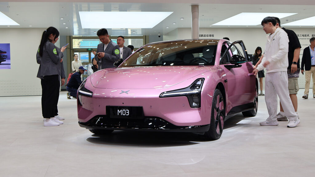 A pink Xpeng Mona M03 electric sedan on display as visitors examine it at the 2026 Beijing auto show.
