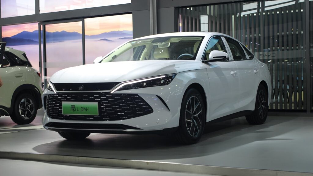 White BYD Qin L DM-i plug-in hybrid sedan on display indoors, viewed from the front and side.
