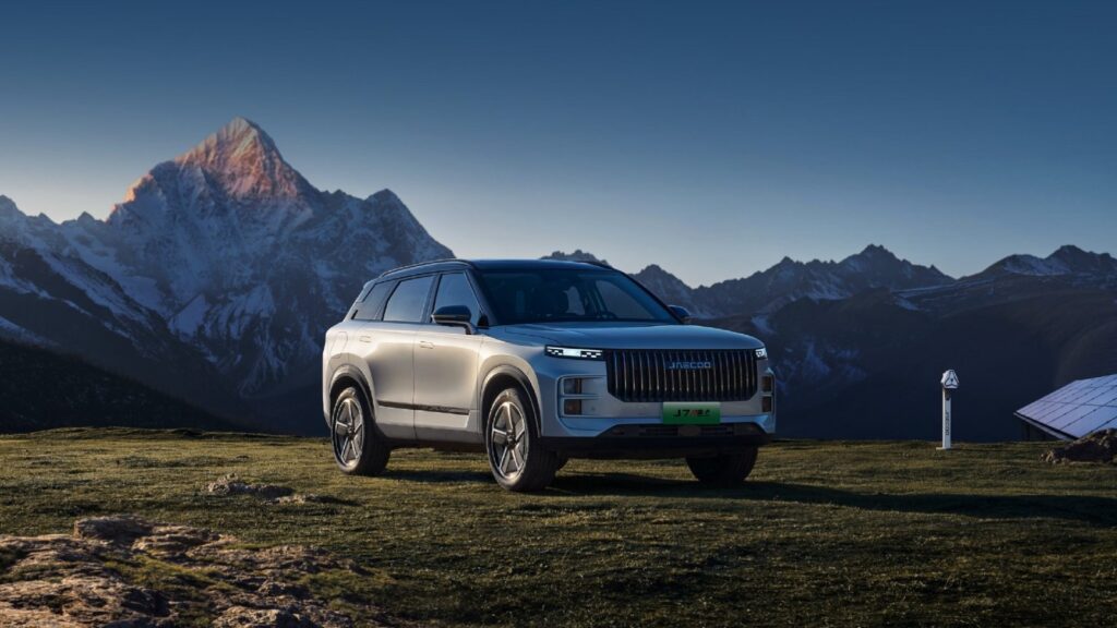 Silver Jaecoo 7 SUV viewed from the front three-quarter angle, parked on grass against a backdrop of snow-covered mountains.