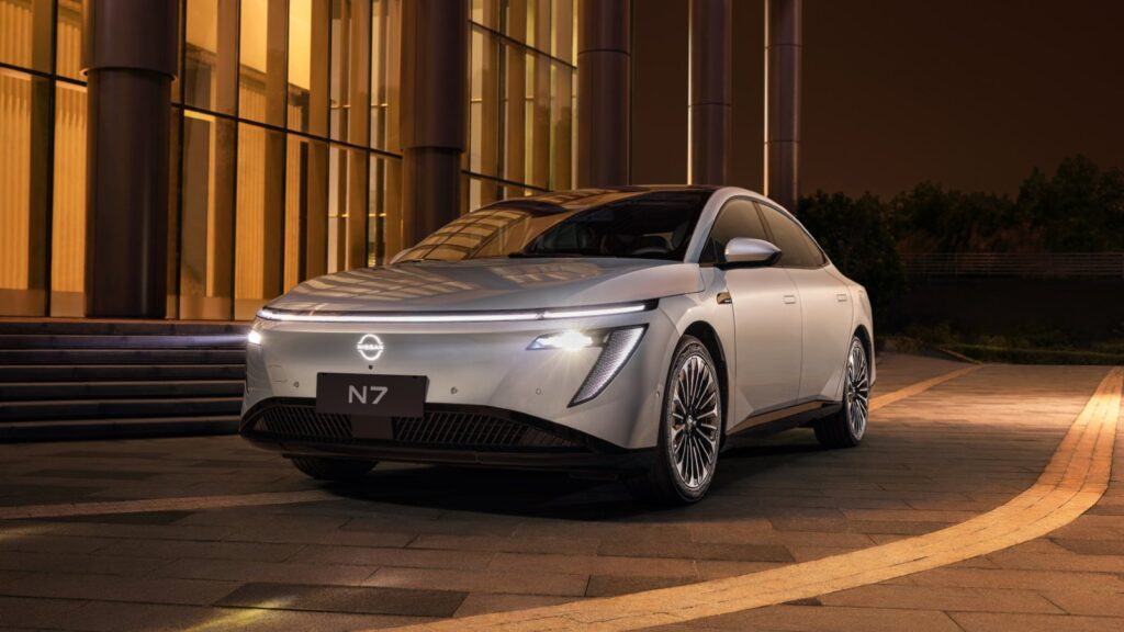 A silver Nissan N7 electric sedan parked at night in front of a modern glass building with warm amber lighting, shown from a front three-quarter angle with its full-width LED light bar and illuminated Nissan badge glowing.