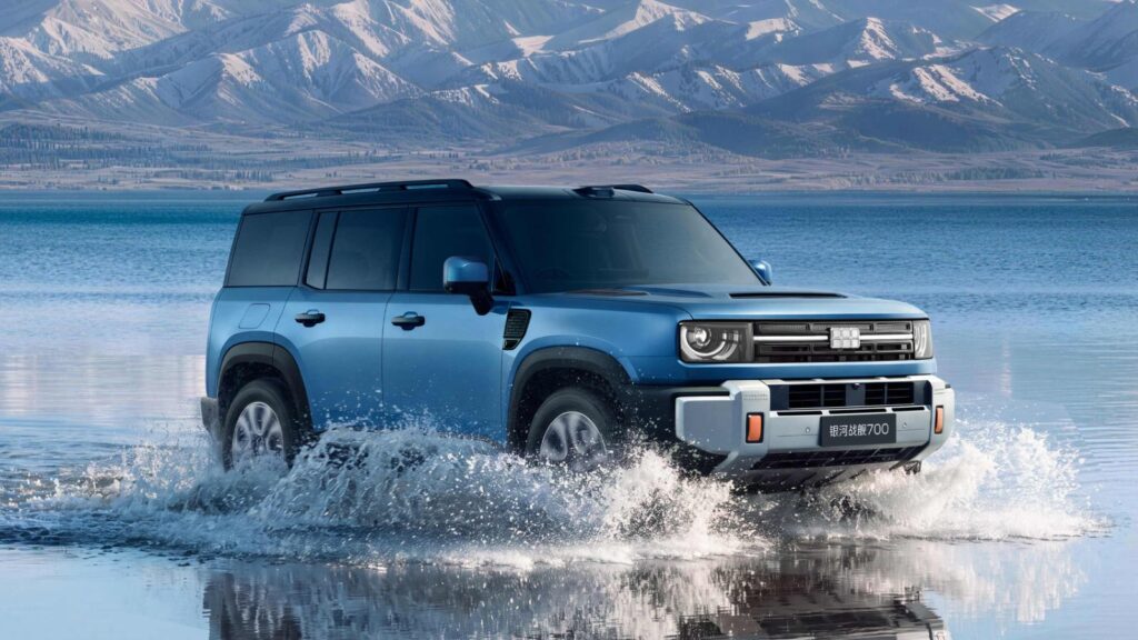Blue Geely Galaxy Cruiser 700 driving through shallow water, with a lake and snow-covered mountains in the background.