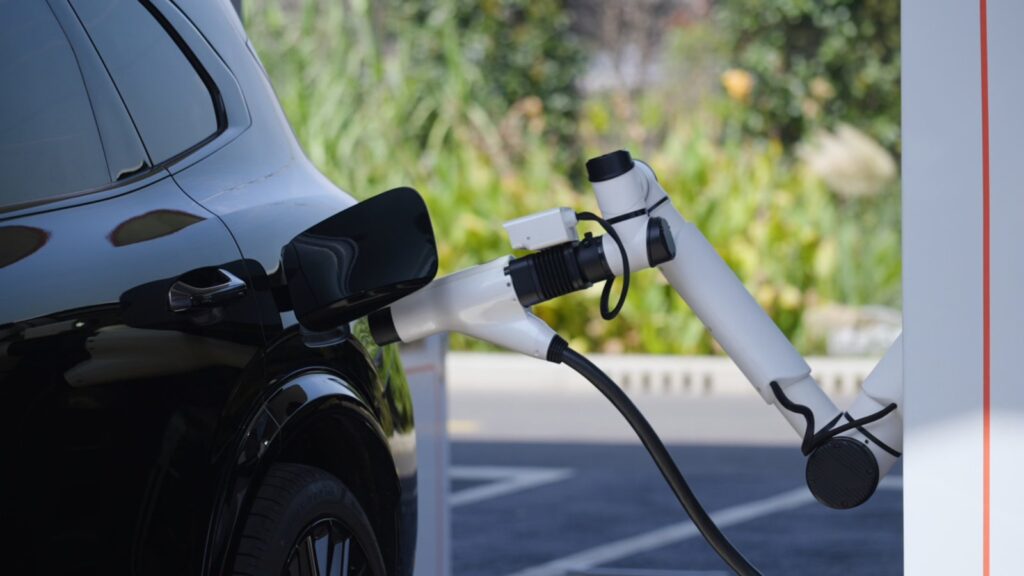 Geely takes on BYD with system charging EVs to 70% in 4.5 minutes A white robotic charging arm plugs a connector into the charging port of a black electric vehicle parked outdoors, with green shrubs and trees in the background.
