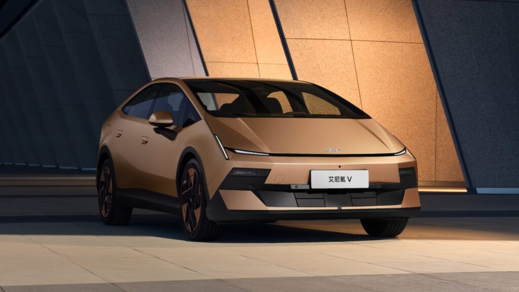 Gold Hyundai Ioniq V electric sedan viewed from the front three-quarter angle outside a modern building.