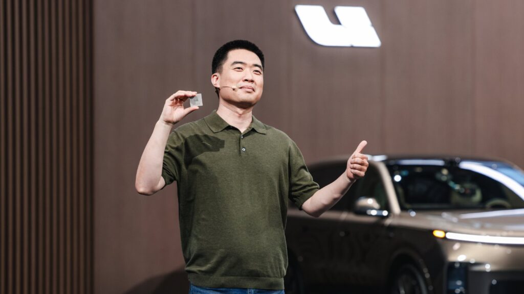 Li Auto founder, chairman and CEO Li Xiang stands on stage holding up the Mach M100 chip between his fingers with one hand and giving a thumbs-up with the other, with a Li Auto logo and a vehicle in the background at Livis Day in June 2026.