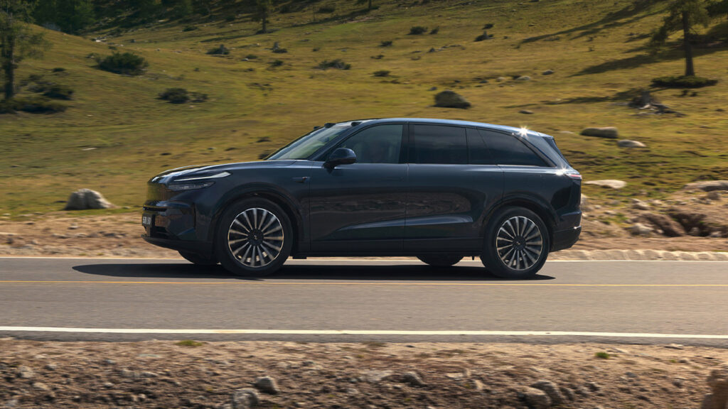 Side view of an Onvo L80 in Xinghan Blue with 21-inch multi-spoke wheels driving past a grassy hillside.