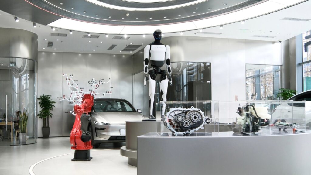 A Tesla Optimus humanoid robot stands on a display platform at a Tesla store in Beijing, next to a red industrial robot arm, a silver Model Y and drivetrain components in glass cases.