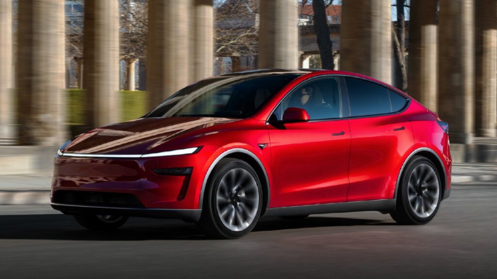 Red Tesla Model Y driving past stone columns, viewed from the front three-quarter angle.