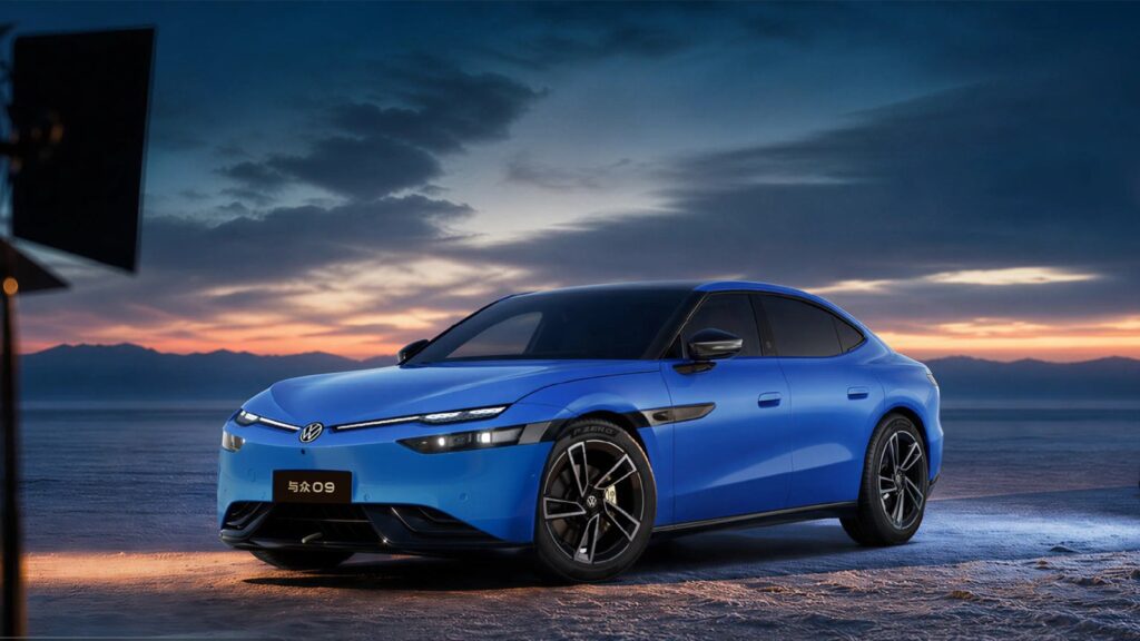 A blue Volkswagen ID. UNYX 09 electric sedan shown from the front three-quarter angle at dusk on a flat open landscape, with a full-width LED light bar, illuminated VW badge and black five-spoke wheels, against a dramatic sunset sky and distant mountains.