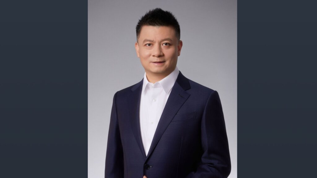 Portrait of Jeff Zhang, incoming president of Audi China, in a dark navy suit and white open-collar shirt against a light gray background.