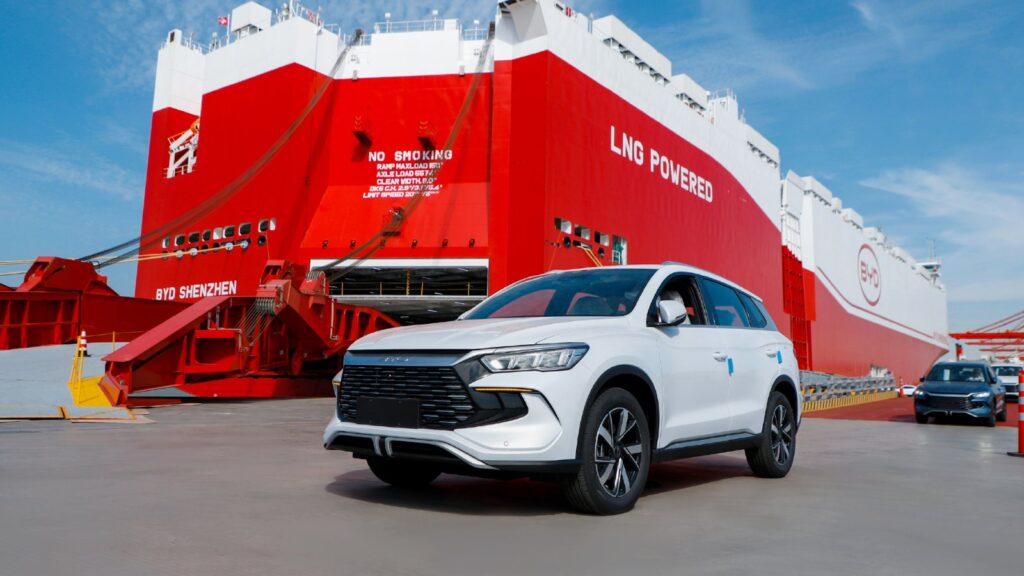 A white BYD SUV beside the red-and-white BYD Shenzhen car carrier at a port.