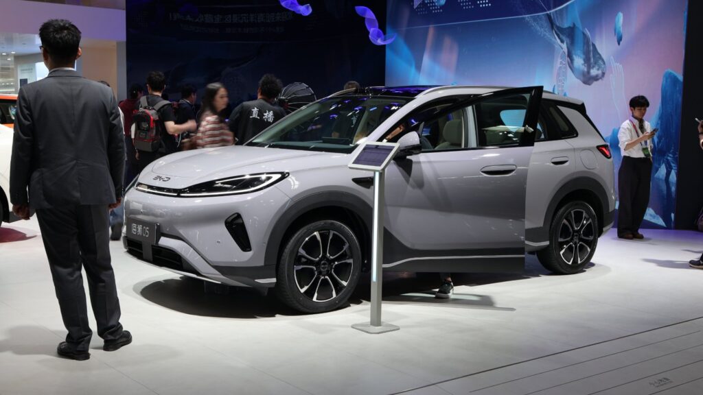A silver BYD Sealion 05 SUV with its front door open on an auto show floor, as attendees walk past and a man in a gray suit stands nearby.