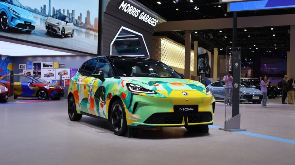 MG4 electric hatchback with a colorful wrap and black roof, viewed from the front three-quarter angle at an auto show.