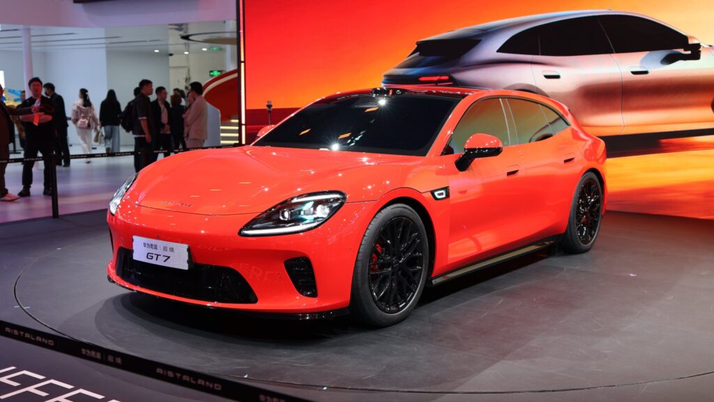 A red Aistaland GT7 shooting brake displayed on a rotating platform at the Beijing auto show, with a large screen behind it showing the same model and visitors walking in the background.