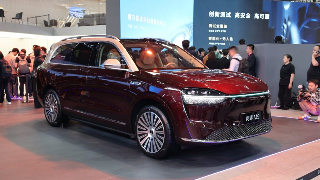 A dark red Aito M9 full-size SUV on display on a turntable at an indoor auto show stand, shown from the front three-quarter angle, with visitors and media gathered around it.