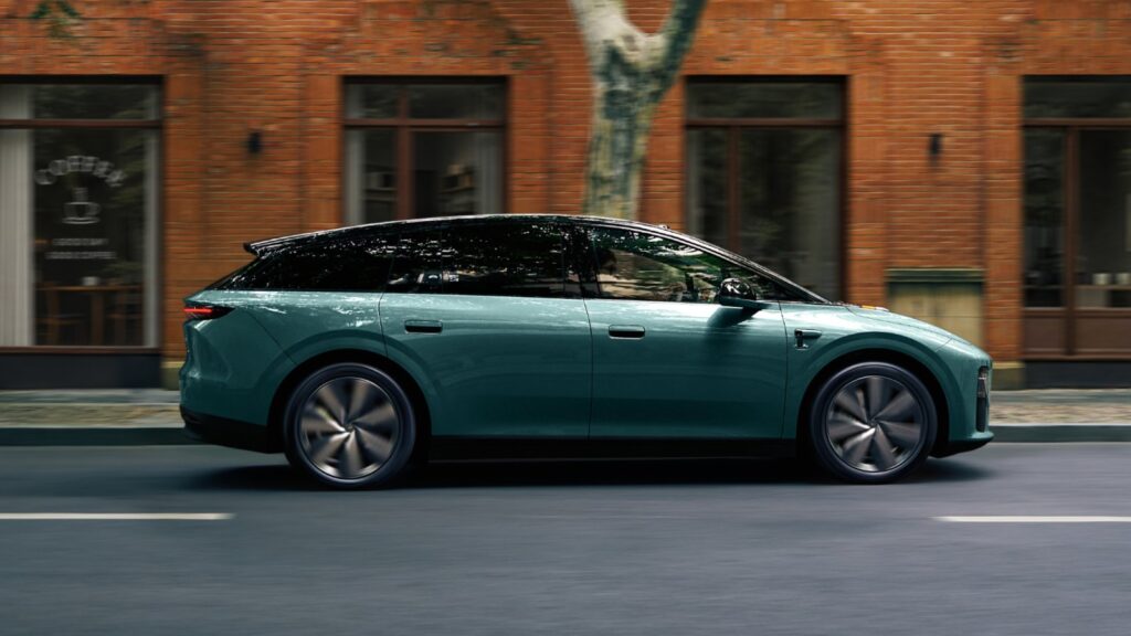A side profile of the 2026 Li i6, a teal-green all-electric SUV, driving along a city street past a red-brick building with a coffee shop and a tree.