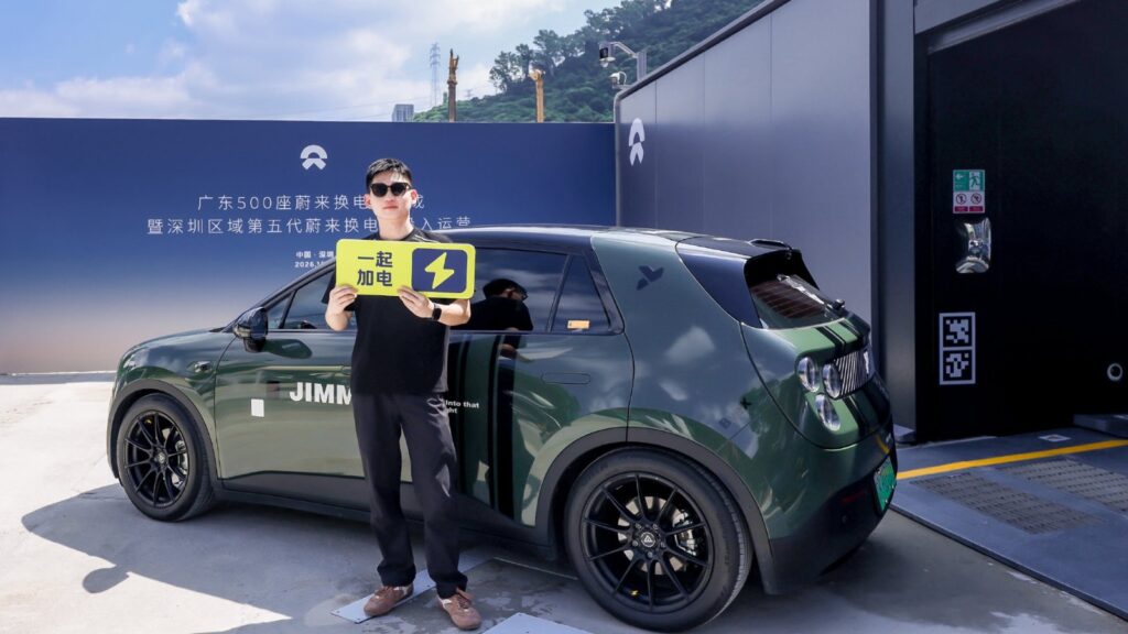 A man holds a yellow charging-themed sign beside a green electric hatchback outside a Nio battery swap station.