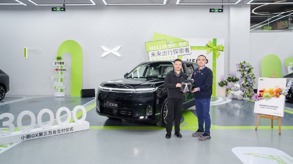 He Xiaopeng, left, and a GX owner hold a commemorative gift beside a black Xpeng GX at a delivery ceremony.