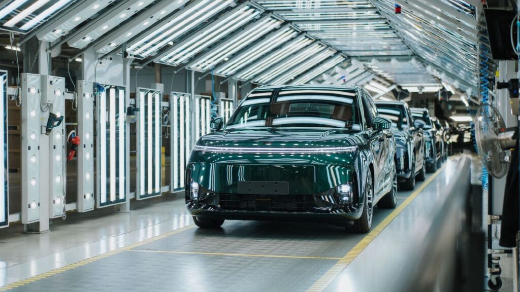 A dark green Xpeng G9L leads a line of vehicles through an illuminated factory inspection area.