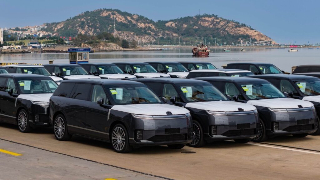 Rows of Xpeng GX SUVs with protective coverings parked beside the water at a port.
